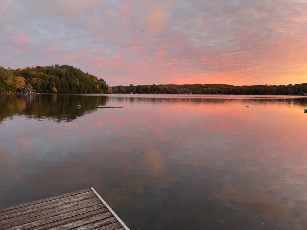 A dock at sunset on a calm Muskoka lake, pink and orange cloud reflected across the water