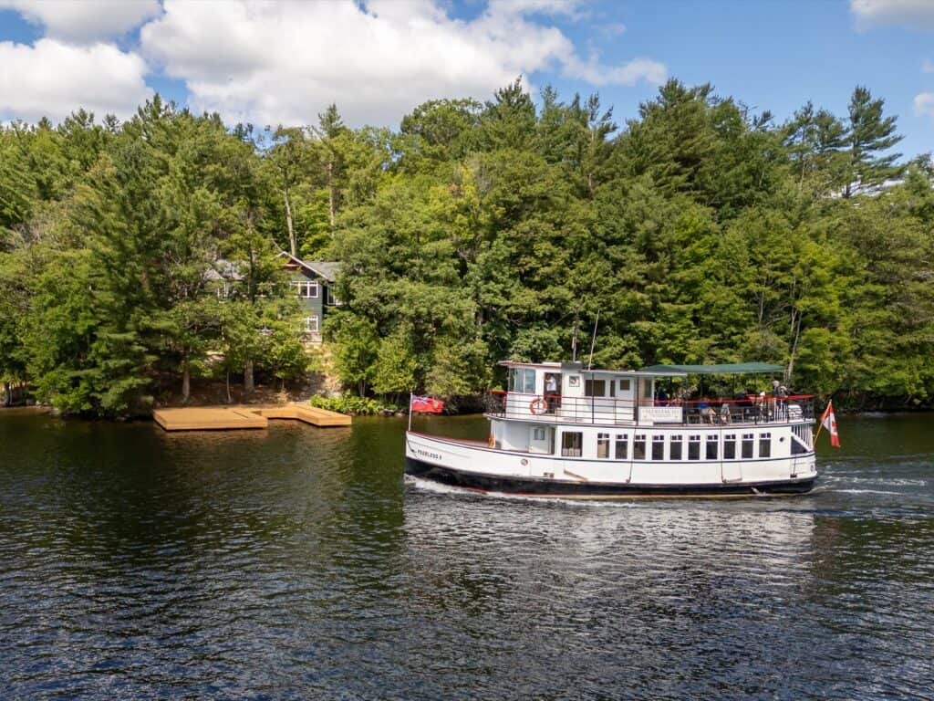 The Peerless cruising past a treed shoreline on Lake Muskoka with a cottage and dock behind her