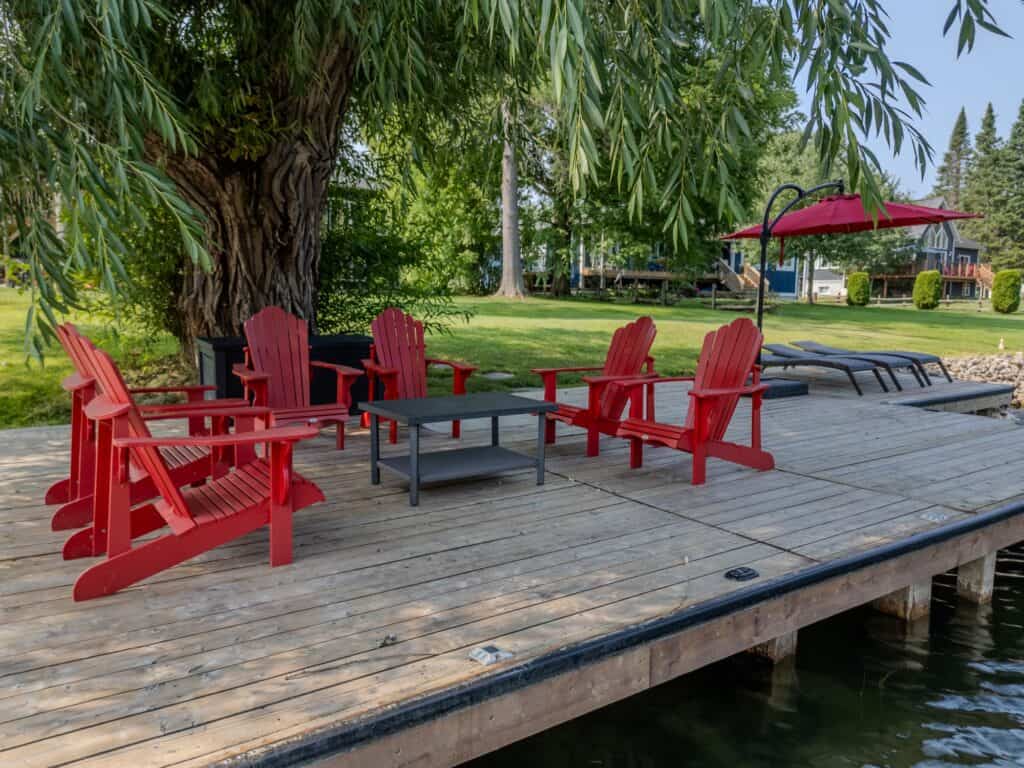 Red Muskoka chairs arranged on a wide wood dock under a willow tree, cottages and lawn behind
